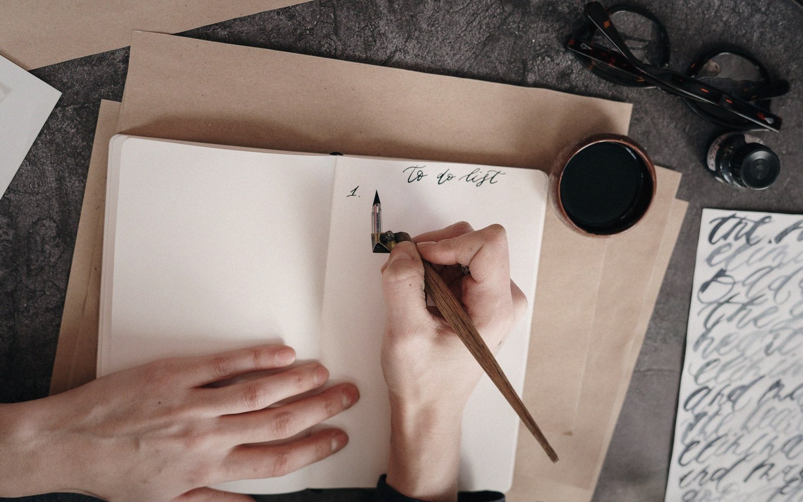 A hand writes "to do list" with a dip pen beside an ink jar and glasses